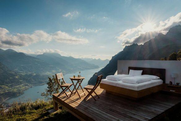 Imagen de una de las habitaciones de Null Stern Hotel, en Suiza. Cama, mesa y sillas sobre una tabla de madera con las montañas suizas y un lago de fondo.